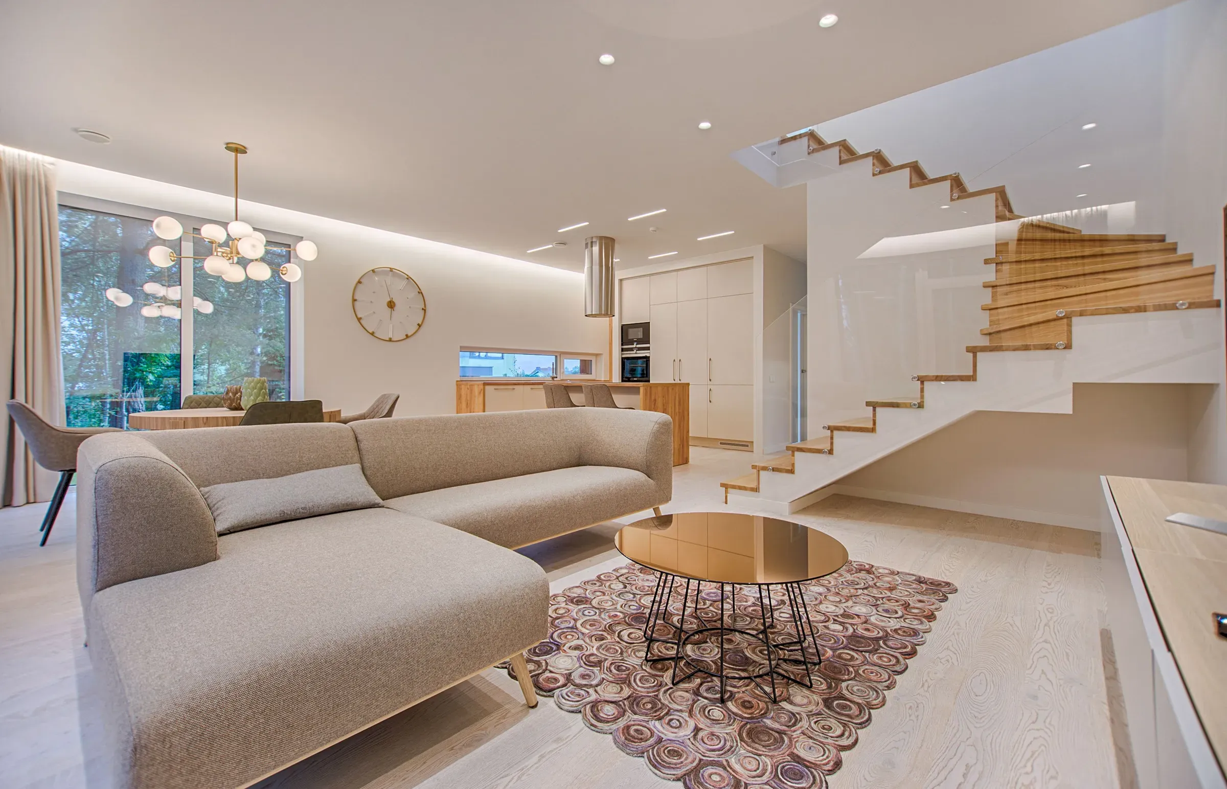 An open-plan living room with a pale sofa, a timber staircase and full-height windows letting in daylight.